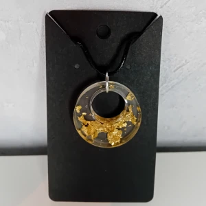 Egentillverkat halsband med guldflingor i cirkelhänge - Egentillverkade halsband gjort i resin med ett cirkelformat hänge fyllt med guldflingor. Hänget är transparent och hänger på en svart rem. Perfekt för att ge en elegant touch till din stil. Cirkeln är 3x3cm stort