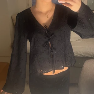 Svart blus med knytning - Snygg svart blus med knytning framtill och broderade detaljer. Blusen har långa ärmar och en v-ringad design. 