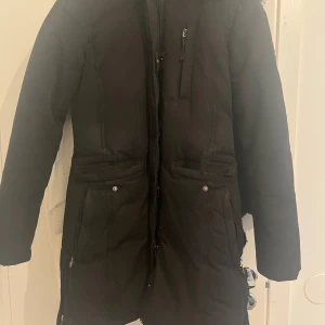 Svart parkajacka från Hollies - Snygg svart parkajacka från Hollies med pälskantad huva. Jackan har dragkedja och knappar framtill samt flera praktiska fickor. Perfekt för kalla dagar. Jackan sälj ej längre och köpt 6000 nypris säljer nu för halva 