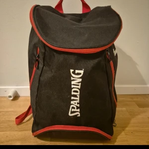 Svart ryggsäck från Spalding  - Säljer en svart ryggsäck från Spalding med röda detaljer och dragkedjor. Perfekt för sport och träning. Ryggsäcken har ett stort huvudfack och flera mindre fack för enkel organisering.