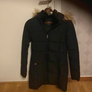 Svart dunjacka från Lindex - Snygg svart dunjacka från Lindex med pälskantad huva. Jackan har en dragkedja framtill och långa ärmar. Perfekt för kyliga dagar.