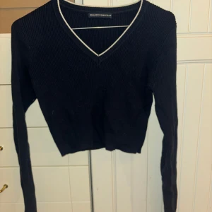 Svart ribbad tröja från Brandy Melville - Säljer en svart ribbad tröja från Brandy Melville med v-ringning och vita detaljer vid halsen. Tröjan har långa ärmar och en croppad passform. Perfekt för en stilren look!