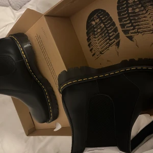 Svarta boots från Dr. Martens HELT NYA - Säljer ett par klassiska svarta Dr. Martens boots med gul söm. De har en slät yta och är perfekta för en stilren look. Skorna har en robust sula och är kända för sin hållbarhet. Skorna är helt nya oanvända