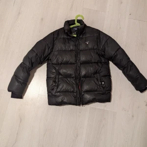 Svart dunjacka från GANT - Säljer en svart dunjacka från GANT i storlek 146/152 cm för åldern 11-12 år. Jackan har en dragkedja framtill och långa ärmar med ribbade muddar. Perfekt för kyliga dagar. Finns två små revor, se bilder.