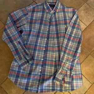 Rutig skjorta från Ralph Lauren - Snygg rutig skjorta från Ralph Lauren i blå, rosa och gröna toner. Skjortan har en slim fit passform och är tillverkad i 100% bomull med stretch. Den har långa ärmar och knappar framtill samt en broderad logotyp på bröstet. Skick 10/10 
