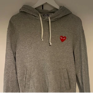  Comme des Garçons hoodie - Stilren grå hoodie från Comme des Garçons med det ikoniska röda hjärtat på bröstet. Tröjan har en justerbar huva med vita snören och praktiska fickor framtill. Nyskick; 10/10. Skriv vid frågor och funderingar!
