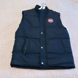 Canada Goose - Säljer en stilren mörkblå dunväst från Canada Goose med broderad logotyp på bröstet. Västen har knappar och dragkedja framtill samt praktiska fickor. Perfekt för alla dagar.