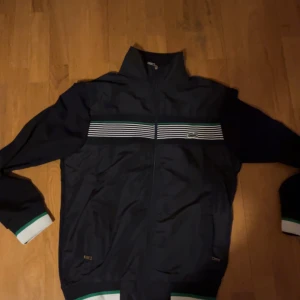 Mörkblå jacka från Lacoste - Säljer en mörkblå jacka från Lacoste med vita och gröna ränder över bröstet. Jackan har dragkedja och broderad logga på ärmen. Perfekt för storlek M elr L
