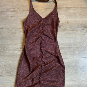 Glittrig brun klänning - Snygg glittrig brun klänning med halterneck och rynkad detalj framtill. Perfekt för en kväll ute med vänner. Klänningen har en åtsittande passform som framhäver figuren.