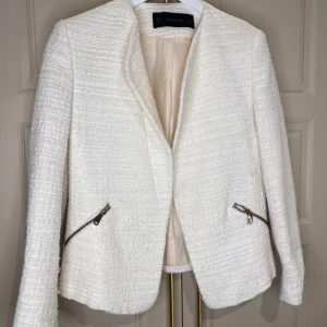 Vit kavaj från Zara Basic - Elegant vit kavaj från Zara Basic med dragkedjedetaljer på fickorna. Tillverkad i ett strukturerat material som ger en stilren look. Perfekt för att lyfta vilken outfit som helst. Storlek M