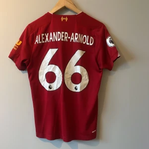 Röd Liverpool fotbollströja från New Balance - Säljer en röd Liverpool fotbollströja med Alexander-Arnold och nummer 66 på ryggen. Tröjan har vita detaljer och Premier League-märke på ärmen. Tillverkad av New Balance med Standard Chartered som sponsorlogga på framsidan.
