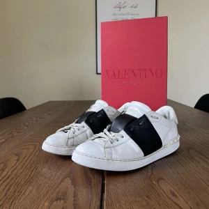 Valentino Garavani sneakers - Snygga Valentino Garavani sneakers i vitt skinn med en bred svart rem över snörningen. Skorna har en klassisk design med rund tå och är perfekta för en stilren look. Skorna är i okej skick hör av dig vid frågor!! 