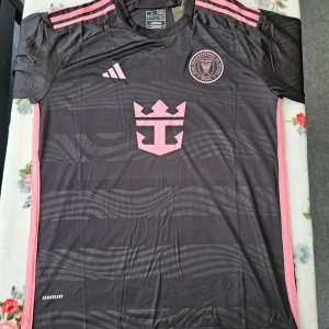Svart och rosa fotbollströja från Adidas - Snygg svart fotbollströja med rosa detaljer från Adidas. Tröjan har korta ärmar och ett stilrent mönster med klubbemblem på bröstet. Perfekt för fotbollsfans som vill sticka ut på planen!