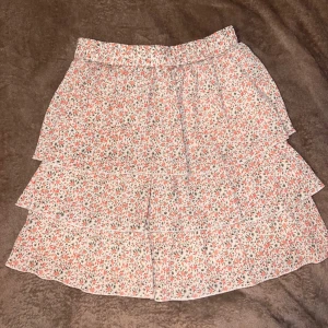 Blommig kjol  - Charmig blommig kjol från Yu & Me i storlek M. Kjolen har ett vackert blommönster i rosa och grönt på en vit bakgrund. Den är designad med volanger som ger en lekfull och feminin touch.