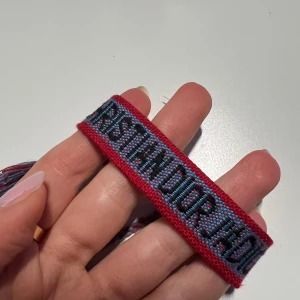 Dior armband - Unikt armband från Christian Dior i rött och blått med dekorativa tofsar. Perfekt för att ge en färgglad touch till din outfit. Bältet har en vävd text och hjärtdetalj som ger en personlig stil.