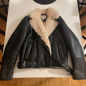 Svart skinnjacka med foder från H&M - Snygg svart skinnjacka från H&M med mjukt pilefoder. Jackan har en dragkedja framtill och ett bälte i midjan för extra stil. Perfekt för kyligare dagar med sin varma och trendiga design. Storlek XS