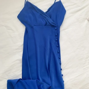 Blå klänning från Zara - Snygg blå klänning från Zara med smala axelband och omlott-design. Klänningen har dekorativa knappar längs sidan och är perfekt för en elegant look.