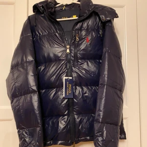 Mörkblå dunjacka från Polo Ralph Lauren - Snygg mörkblå dunjacka från Polo Ralph Lauren med dragkedja och huva. Jackan har en broderad logga på bröstet och praktiska fickor med dragkedja. Perfekt för kyliga dagar. 1|1