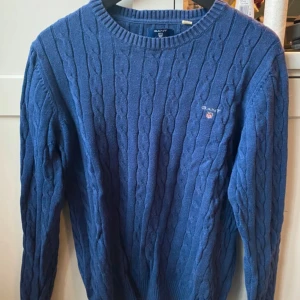 Blå kabelstickad tröja från GANT - Snygg blå kabelstickad tröja från GANT med rund halsringning och broderad logotyp på bröstet. Perfekt för kyligare dagar. Skriv vid eventuella frågor.