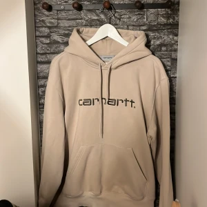 Beige hoodie från Carhartt - Säljer en stilren beige hoodie från Carhartt med klassisk logga på bröstet. Tröjan har en rymlig huva med dragsko och en praktisk magficka. Perfekt för en avslappnad stil.