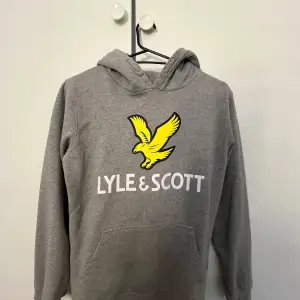 Säljer en snygg grå hoodie från Lyle & Scott i mycket bra skick. Den har en stor gul logga på bröstet och en praktisk magficka. Perfekt för både skola och fritid. Passar ungdomar i åldern 14-15 år.