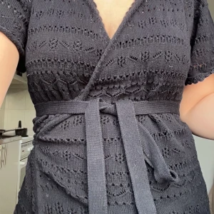 Svart omlottklänning med knytband - Snygg svart omlotttröja i stickat material med v-ringning och korta ärmar. Den har ett fint knytband i midjan som ger en smickrande passform. Perfekt för både vardag och festliga tillfällen!