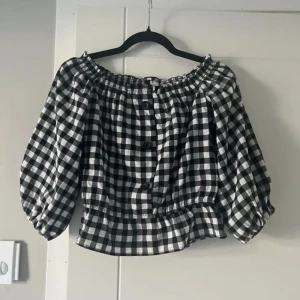 Svartvit rutig offshoulder topp - Säljer en superfin svartvit rutig offshoulder topp med knappar framtill. Den har en elastisk kant upptill och volangdetaljer vid midjan. 