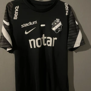 Svart fotbollströja från Nike - Säljer en svart fotbollströja från Nike med AIK-logga på bröstet. Tröjan har korta ärmar med grå och vita detaljer. Sponsorer som 'notar' och 'Stadium' är tryckta på framsidan. Perfekt för fotbollsfans! ⚽️