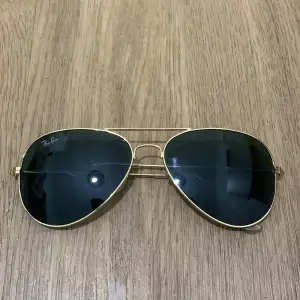 Säljer ett par klassiska Ray-Ban solglasögon i pilotstil med guldfärgad metallram och mörka linser. Perfekta för en stilren look i sommar. De är i nytt skick och redo att användas för att skydda dina ögon med stil. Passar både till vardags och fest! Köpte den för 1299. Helt äkta!