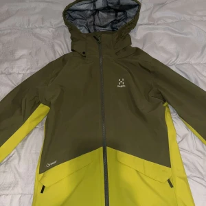 Grön och gul jacka från Haglöfs - Säljer en grön och gul jacka från Haglöfs i mycket bra skick. Den har en praktisk huva och dragkedja framtill. Perfekt för utomhusaktiviteter och håller dig torr och bekväm. Jackan är långärmad och har en snygg design med kontrasterande färger.