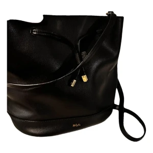 Svart axelväska från Ralph Lauren - Snygg och stilren svart axelväska från Ralph Lauren i skinn. Väskan har en praktisk dragsko och eleganta gulddetaljer. Perfekt för både vardag och fest! 🖤I nyskick knappt använd‼️Nypris: 5 999kr 