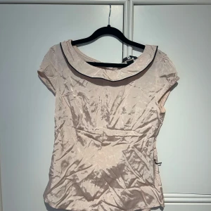 Ljusrosa blus med svart kant - Säljer en elegant beige blus med svart kant runt halsen. Den har korta ärmar och är gjord i ett glansigt material som ger en lyxig känsla. Perfekt för både vardag och fest! Den har haft ett skärp i midjan därav de svarta trådarna på bild 4 med de går ju att klippa bort! Lappen säger 40 men skulle säga att den passar XS-M!