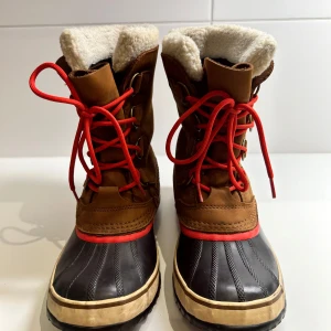 Vattentäta vinterstövlar från Sorel - Säljer ett par robusta och varma vinterstövlar från Sorel i bra skick, storlek 40. De är bruna med svarta detaljer och har rosa snören. Insidan är fodrad med mjukt material för extra värme. Perfekta för kalla och blöta vinterdagar. Sulan är mönstrad för bra grepp. 