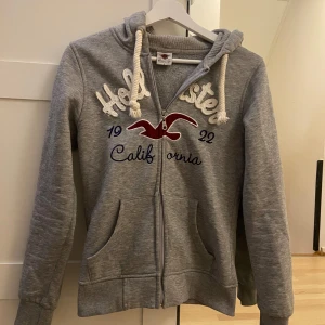 Grå hoodie från Hollister - Säljer en grå hoodie från Hollister med dragkedja och stor logga på framsidan. Den har en mysig huva med vita snören och praktiska fickor. Perfekt för en avslappnad stil eller kyliga dagar. 💞