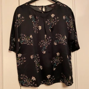 Svart blommig blus från Ellos - Snygg svart blus från Ellos med ett vackert blommönster i olika färger. Blusen har korta ärmar och en rund halsringning med en liten knappdetalj i nacken. Perfekt för både vardag och fest! Oanvänd🤩
