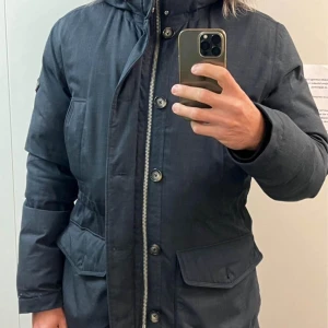 Mörkblå parkajacka med huva - Säljer en snygg mörkblå parkajacka med huva. Jackan har dragkedja och knappar framtill samt praktiska fickor. Perfekt för höst och vinter, håller dig varm och stilren. Passar både till vardags och lite finare tillfällen.