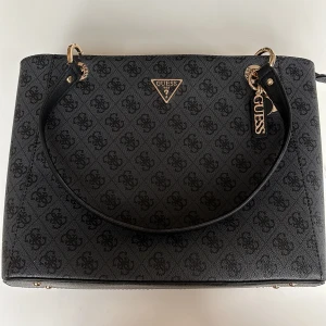 Svart axelväska från Guess - Snygg oanvänd svart väska från Guess med deras ikoniska logotypmönster. Väskan har guldfärgade detaljer och en praktisk dragkedja upptill. Perfekt för både vardag och fest! 🖤