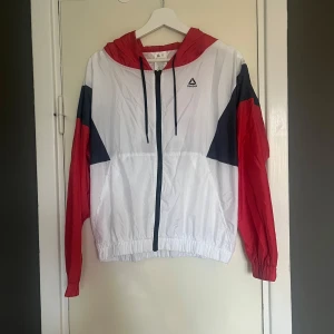 Vit och röd vindjacka från Reebok - Säljer en snygg vindjacka från Reebok i vitt med röda och mörkblå detaljer. Jackan har en huva och dragkedja framtill. Perfekt för vår och höst, och den har en sportig look som funkar både till träning och vardag. Stor Reebok-logga på ryggen ger den en cool retrostil.