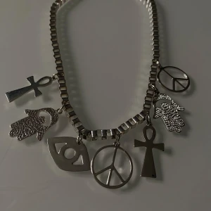 Silverfärgat halsband med symboler - Coolt silverfärgat halsband med flera hängande symboler, inklusive fredstecken, ankh och hamsa. Perfekt för att ge din outfit en unik touch. Kedjan är robust och hängena är detaljerade, vilket gör det till ett statement-smycke.
