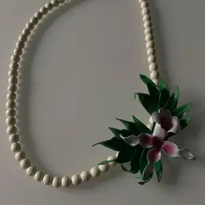 Säljer ett unikt pärlhalsband med en vacker blomdetalj i rosa och grönt. Halsbandet har en klassisk design med vita pärlor och en iögonfallande blomma som ger en tropisk känsla. Perfekt för att liva upp vilken outfit som helst!