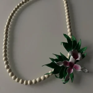 Pärlhalsband med blomdetalj - Säljer ett unikt pärlhalsband med en vacker blomdetalj i rosa och grönt. Halsbandet har en klassisk design med vita pärlor och en iögonfallande blomma som ger en tropisk känsla. Perfekt för att liva upp vilken outfit som helst!