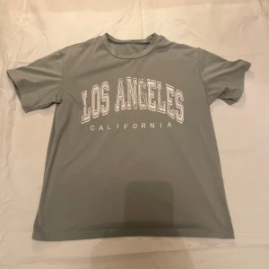 Grön t-shirt med Los Angeles-tryck - Säljer en grön t-shirt med trycket 'Los Angeles California' i vitt. Perfekt för en avslappnad stil och passar bra till jeans eller shorts. T-shirten är kortärmad och i bomull, vilket gör den skön att bära under varma dagar.