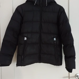 Svart dunjacka från McKinley - Säljer en svart dunjacka från McKinley i storlek 146/152. Modell: Terry jr                                   Jackan är mycket varm och bekväm med sitt vadderade material i 100% polyester. Den har en dragkedja framtill och två praktiska fickor, varav ena dragkedjan är trasig (se bild 5). Huvan ger extra skydd mot väder och vind. Perfekt för vinter och höst!🌟