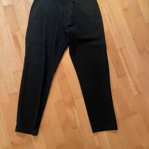 Svarta kostymbyxor - Säljer ett par stilrena svarta kostymbyxor i bra skick. De har en klassisk passform med knappar bak och bälteshällor. Perfekta för både formella tillfällen och vardagsbruk. Passar till alla säsonger och kan enkelt matchas med olika överdelar.