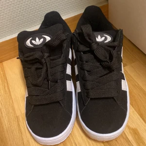 Svarta Adidas Campus sneakers - Säljer ett par snygga svarta Adidas Campus sneakers i mycket bra skick. De har de klassiska vita ränderna på sidan och en vit sula. Perfekta för både vardag och fest! Skorna har snörning och är gjorda i syntetmaterial.