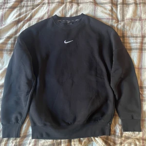 Svart sweatshirt från Nike - Säljer en klassisk svart sweatshirt från Nike i mycket bra skick. Den har en enkel design med en liten vit Nike-logga på bröstet. Perfekt för både träning och vardag. Mjuk och bekväm med ribbade muddar vid ärmslut och nederkant.