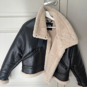 Vinterjacka  med foder från Zara - Säljer en snygg svart skinnjacka från Zara med mysigt beige foder. Jackan har en cool, croppad stil och är perfekt för höst och vinter. Den har långa ärmar och en stilren design med dragkedja och spänne vid kragen. Perfekt för att hålla sig varm och trendig! 🖤