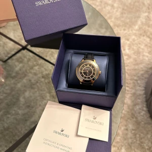 Swarovski klocka i guld och svart - Säljer en elegant Swarovski klocka med en stilren design. Klockan har en svart urtavla med guldiga detaljer och ett svart läderarmband. Perfekt för både vardag och fest! Kommer i originalförpackning med certifikat och instruktioner.
