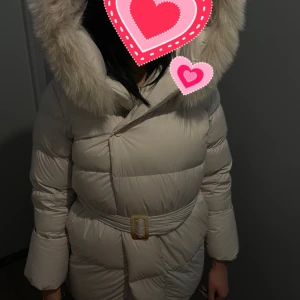 Beige dunjacka med pälshuva - Säljer en supermysig beige dunjacka med stor pälshuva. Jackan har ett bälte i midjan för en snygg passform och knappar framtill. Perfekt för kalla vinterdagar! 🧥❄️