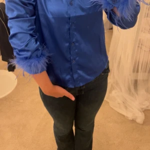 Blå och svart satinblus med fjäderdetaljer - Säljer två snygga blusar i satin, en i blått och en i svart, med coola fjäderdetaljer vid ärmsluten. Perfekt för en festlig kväll! Blusarna har knappar framtill och en elegant glansig finish.